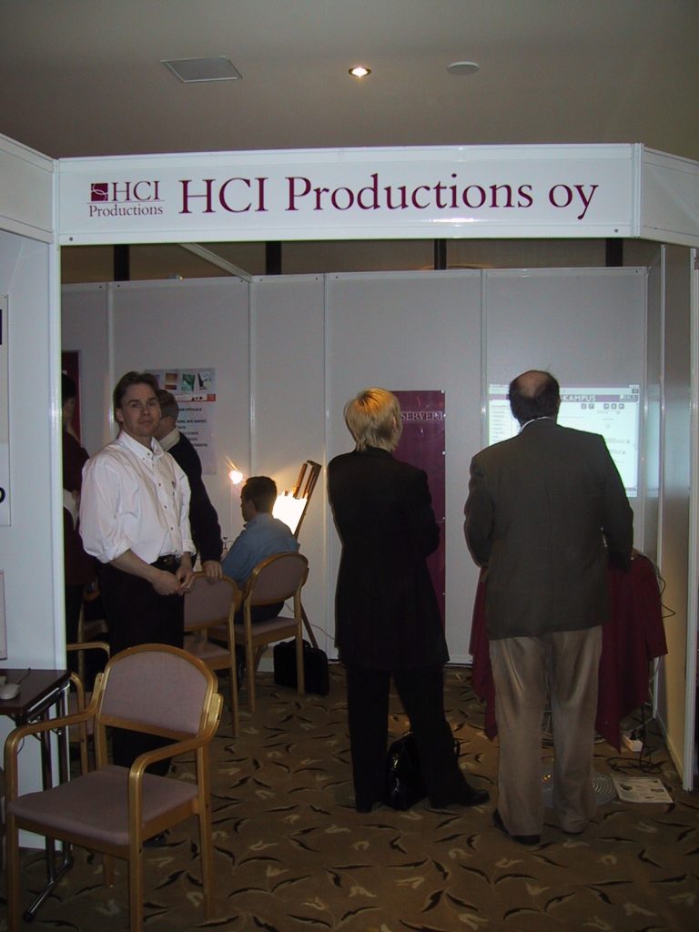 HCI Productions oy:n messu ständi konferenssissa. Ihmisiä seisomassa ständillä.